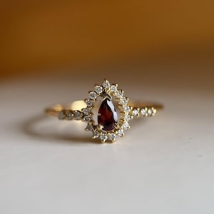 Puede incluir: Un anillo de oro con una piedra preciosa granate de color rojo intenso en forma de pera, rodeada por un halo de pequeños diamantes blancos de talla redonda. El aro es fino y metálico, con diamantes adicionales engastados en los hombros.