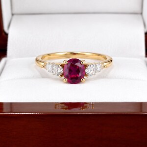 Op de afbeelding: Een gouden ring met een centrale ovaal geslepen dieprode robijn, geflankeerd door twee kleine rond geslepen heldere diamanten aan elke kant. De ring wordt gepresenteerd in een gepolijst donkerhouten doosje met een witte binnenvoering.
