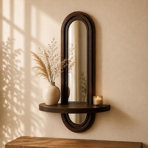 Puede incluir: Un espejo de pared de madera oscura con un marco ovalado alargado y un estante semicircular integrado. Un jarrón de cerámica color crema con hierbas secas y un pequeño portavelas de cristal con una vela encendida descansan sobre el estante.