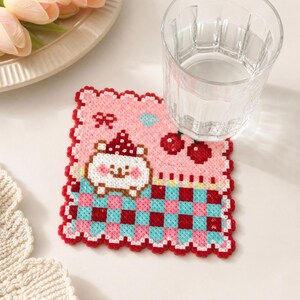 May include: A square coaster made of fused plastic beads featuring a white bear character with a red hat, a pink background with a small blue heart, two red cherries, and a red, pink, and light blue checkered pattern along the bottom edge.