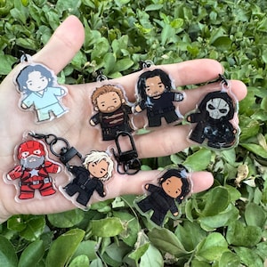 May include: A collection of seven acrylic keychains featuring chibi-style character illustrations. The designs include figures in various outfits, such as a red and blue superhero suit, black tactical gear, and a masked character in a dark hooded cloak.