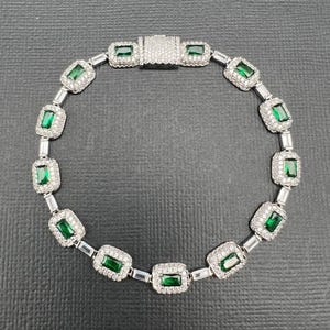 May include: A silver-toned bracelet featuring a series of rectangular emerald-green stones, each surrounded by a halo of small clear stones. The links are connected by small clear stones, and the piece includes a rectangular clasp with clear stones.