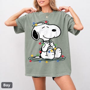 以下が含まれることがあります： セージグリーンの半袖Tシャツ。カラフルなクリスマスライトのコードに絡まった漫画キャラクターのスヌーピーのグラフィックが描かれています。黒い耳を持つ白い犬が座っており、赤、青、黄色、緑の電球に囲まれているデザインです。