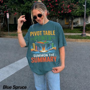 May include: A blue spruce colored short-sleeve t-shirt featuring a graphic print with the text PIVOT TABLE POWER SUMMON THE SUMMARY. The design includes a floating spreadsheet grid, colorful bar charts, and stylized light rays on a dark background.