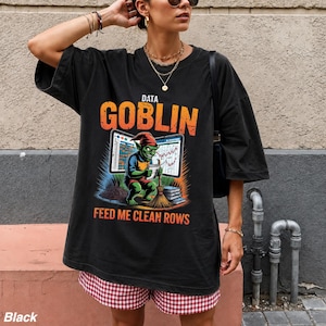 May include: A black oversized t-shirt featuring a graphic print of a green goblin holding a broom in front of computer screens displaying data charts. The text reads DATA GOBLIN at the top and FEED ME CLEAN ROWS at the bottom in orange letters.