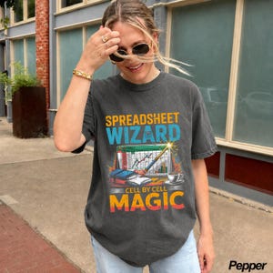 May include: A charcoal gray short-sleeve t-shirt featuring a graphic print with the text SPREADSHEET WIZARD and CELL BY CELL MAGIC. The design includes a computer screen showing a spreadsheet, a magic wand, a stack of books, and a coffee cup.