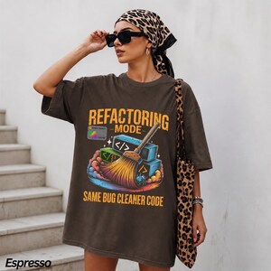 May include: A dark brown oversized t-shirt featuring a graphic print with the text "REFACTORING MODE" and "SAME BUG CLEANER CODE". The design includes a broom sweeping colorful blocks with code symbols and a small blue insect illustration.