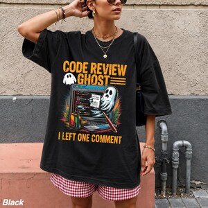 May include: A black short-sleeved t-shirt featuring a graphic print with the text "CODE REVIEW GHOST" and "I LEFT ONE COMMENT". The design includes a cartoon ghost, a laptop screen with code, a magnifying glass, and a pen in vibrant colors.