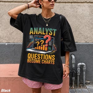 May include: A black oversized t-shirt featuring a graphic print of a laptop displaying a bar chart and a large red question mark. The text reads ANALYST, ANALYST ENERGY, and QUESTIONS BECOME CHARTS in yellow and orange lettering with a retro design.