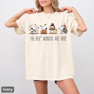 以下が含まれることがあります： アイボリーの半袖Tシャツ。スヌーピーが四季折々のシーンに登場するグラフィックと「THE BER" MONTHS ARE HERE」の文字がプリントされています。イラストには秋の葉、カボチャ、七面鳥、クリスマスツリーと一緒のスヌーピーが描かれています。