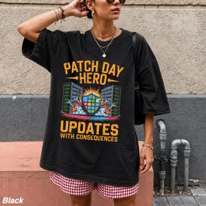 May include: A black oversized t-shirt featuring a graphic print with the text PATCH DAY HERO and UPDATES WITH CONSEQUENCES. The design includes two server towers, a central shield icon with colorful squares, and wrenches on a dark background.