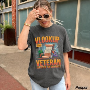 May include: A charcoal gray short-sleeve t-shirt featuring a graphic print with the text VLOOKUP VETERAN SURVIVED THE OLD WAYS. The design includes a scroll, a sword, and a green book, with a grid background and orange and yellow accents.