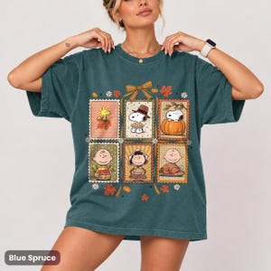 以下が含まれることがあります： ダークティールカラーの半袖Tシャツ。感謝祭を祝うピーナッツのキャラクターたちが描かれた、切手風の長方形のイラスト6枚がプリントされており、秋の葉、カボチャ、中央のリボンで装飾されています。