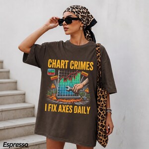 May include: A dark brown oversized t-shirt featuring a graphic print with the text "CHART CRIMES" and "I FIX AXES DAILY". The design depicts a hand holding a wrench against a colorful bar chart, surrounded by warning signs and fire imagery.