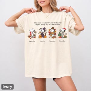 以下が含まれることがあります： アイボリーの半袖Tシャツ。September、October、November、Decemberとラベル付けされた4つの季節のシーンにスヌーピーが描かれています。テキストは、The most wonderful time of the year begins Bring on he ber months と書かれています。