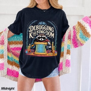 May include: A black t-shirt featuring a graphic of a raccoon wearing armor and reading a scroll at a table with candles. The text reads "DEBUGGING THE KINGDOM" in a stylized font. The shirt is worn under a multi-colored striped knit cardigan.