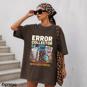 May include: An espresso-colored short-sleeve t-shirt featuring a graphic print with the text ERROR COLLECTOR and GOTTA CATCH THEM ALL. The design includes a glass jar filled with colorful marbles, a magnifying glass, and test report documents.