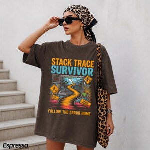 May include: An espresso-colored oversized t-shirt featuring a graphic print with the text STACK TRACE SURVIVOR and FOLLOW THE ERROR HOME. The design includes a computer screen displaying a winding road, yellow warning signs, and colorful foliage.