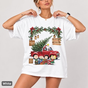以下が含まれることがあります： クリスマスツリーを積んだ赤いピックアップトラックを運転するスヌーピーのグラフィックが描かれた白いTシャツ。ピーナッツのキャラクター、クリスマスの装飾、Christmas Trees、Hot Cocoa、Joy Peace Loveなどの文字が書かれた木製の看板がデザインされています。