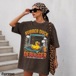 May include: A dark brown oversized t-shirt featuring a graphic of a yellow rubber duck wearing glasses sitting on a laptop. The text reads "RUBBER DUCK DEBUGGER" and "THE DUCK KNOWS EVERYTHING" with a coffee mug nearby. The shirt is paired with a leopard print headscarf.