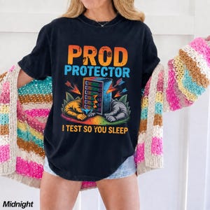 May include: A black short-sleeve t-shirt featuring a graphic print with the text PROD PROTECTOR and I TEST SO YOU SLEEP. The design includes a central server tower, two hands reaching toward it, arrows, and colorful foliage in orange, blue, and green.
