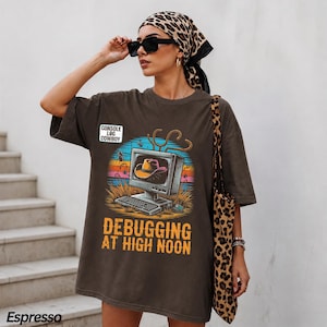 May include: A dark brown short-sleeve t-shirt featuring a graphic of a computer monitor displaying a cowboy hat. The text reads CONSOLE LOG COWBOY and DEBUGGING AT HIGH NOON. The design includes a sunset background, grass, and a lasso.
