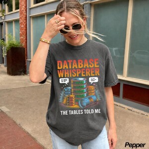 May include: A charcoal gray short-sleeve t-shirt featuring a graphic print with the text "DATABASE WHISPERER" and "THE TABLES TOLD ME". The design includes a stack of colorful cylindrical database icons and speech bubbles on a dark background.