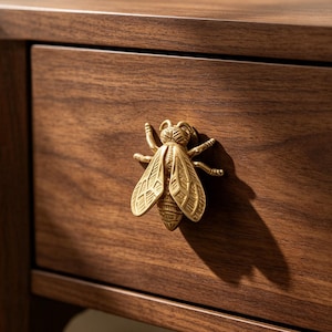 Peut inclure: Un bouton de tiroir en métal doré en forme d'abeille avec des détails texturés sur les ailes et le corps, fixé sur une façade de tiroir en bois brun foncé. La figurine d'abeille présente un abdomen, un thorax et une tête segmentés.