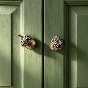 May include: Two metallic acorn-shaped cabinet knobs with a bronze finish attached to sage green wooden cabinet doors. The knobs feature detailed textured caps and smooth bodies, providing a functional grip for opening the cabinet doors.
