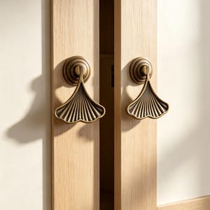 May include: Two antique brass cabinet pulls shaped like ginkgo leaves mounted on light-colored vertical wooden panels. The metal handles feature a fan-like ribbed texture and a circular base, casting distinct shadows on the wood surface.