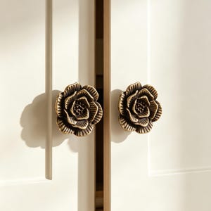 May include: Two antique brass-toned metal cabinet knobs shaped like blooming flowers with layered petals. The knobs are mounted on two white cabinet doors with vertical panel detailing, featuring a dark center and lighter edges.