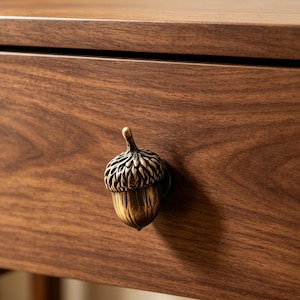 May include: A brass acorn-shaped drawer knob with a textured cap and smooth base, attached to a dark brown wooden drawer front. The metal hardware features a metallic finish with dark recessed details highlighting the natural acorn texture.