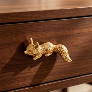 May include: A gold-toned metal drawer pull shaped like a fox in a running pose, attached to a dark brown wooden drawer front. The fox figure features detailed fur textures, pointed ears, and a long, curved tail extending to the right side.
