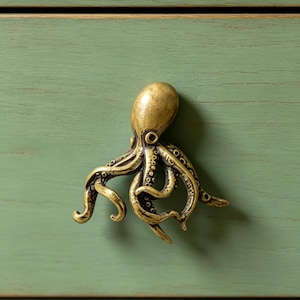 Puede incluir: Un tirador de mueble de metal en tono latón con forma de pulpo, que presenta tentáculos detallados y una cabeza lisa. La figura del pulpo está montada sobre un frente de cajón de madera verde salvia con veta horizontal y acabado desgastado.
