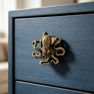 Puede incluir: Un tirador de mueble de metal color latón con forma de pulpo, con tentáculos detallados y una cabeza texturizada. El tirador está montado en un frente de cajón de madera azul marino oscuro con veta visible y un diseño de marco horizontal.