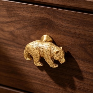 Pode incluir: Um puxador de móvel em metal dourado com formato de urso a caminhar com textura de pelo detalhada. O urso está fixado a uma base circular e montado numa frente de gaveta de madeira castanha escura com um padrão de veios horizontais.