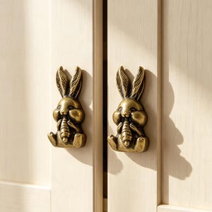 May include: Two antique brass-toned metal cabinet knobs shaped like seated rabbits holding carrots. Each knob features a detailed, three-dimensional rabbit figure with long ears and a textured finish, mounted onto light-colored wooden cabinet doors.