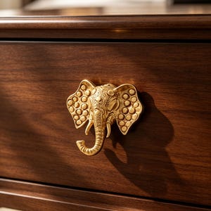 Puede incluir: Un pomo de mueble de metal dorado con forma de cabeza de elefante, con orejas texturizadas y una trompa curvada, montado en un frente de cajón de madera marrón oscuro. El elefante presenta detalles en relieve en la frente y las orejas.