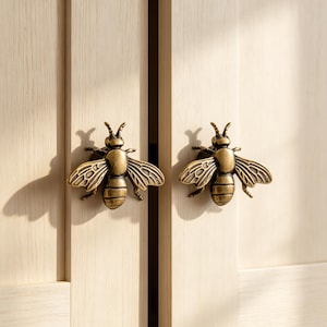 Könnte beinhalten: Zwei Schrankknöpfe aus antikfarbenem Messing in Bienenform mit detaillierten Flügeln und segmentierten Körpern. Diese dekorativen Beschläge sind an hellen Holzschranktüren mit vertikaler Maserung befestigt.