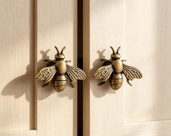 May include: Two antique brass-toned metal cabinet knobs shaped like bees, featuring detailed wings and segmented bodies. These decorative hardware pieces are mounted onto light-colored wooden cabinet doors with vertical grain patterns.