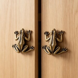 Puede incluir: Dos pomos de armario de metal en tono latón con forma de rana, cada uno con extremidades detalladas y un dorso texturizado, montados en dos puertas de armario de madera clara con un patrón de veta de madera vertical visible.