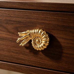 May include: A gold-toned cabinet knob shaped like a nautilus shell with extended tentacles, mounted onto a dark brown wooden drawer front. The metallic finish is reflective and smooth, highlighting the spiral ridges and detailed texture of the shell.