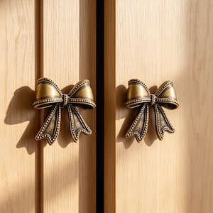 May include: Two antique brass-toned metal cabinet knobs shaped like bows with beaded edges and a dark, weathered finish. These decorative hardware pieces are mounted on light-colored wooden cabinet doors with vertical grain patterns.