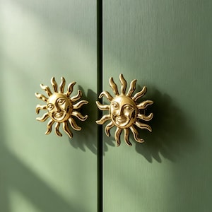 May include: Two polished gold-toned metal cabinet knobs shaped like suns with smiling faces and radiating wavy rays. The knobs are mounted on a flat, matte sage green surface with a vertical seam running between the two circular sun designs.