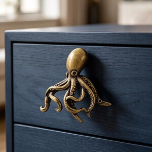 Puede incluir: Un tirador de cajón de metal en tono latón con forma de pulpo fijado a un cajón de madera azul marino oscuro. La figura del pulpo presenta una cabeza redondeada y lisa y varios tentáculos texturizados que sirven como asa funcional.