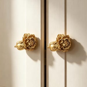 May include: Two gold-colored cabinet knobs shaped like ruffled flowers or cabbages, each featuring intricate, layered petal-like textures. These metallic knobs are mounted on a pair of light-colored, vertical-paneled cabinet doors with a smooth finish.