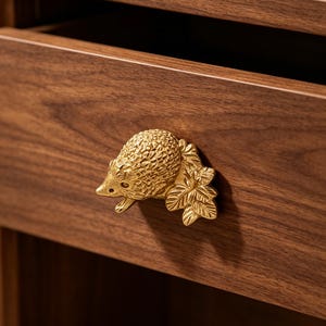 Puede incluir: Un pomo de mueble de metal dorado con forma de erizo junto a un grupo de hojas. La figura animal texturizada y tridimensional está fijada en la parte frontal de un cajón de madera marrón oscuro, funcionando como un tirador práctico.