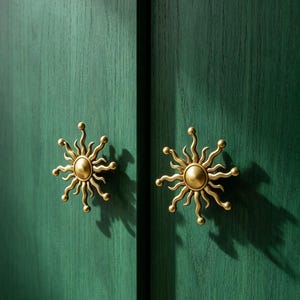 May include: Two brass-colored metal cabinet knobs shaped like suns with radiating wavy rays and a central circular dome. These decorative hardware pieces are mounted on a dark green wooden cabinet door surface with a visible vertical wood grain texture.