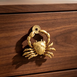 May include: A gold-colored metal drawer pull shaped like a crab holding a circular coin with a square hole in the center. The crab has detailed legs and a textured shell, mounted onto a dark brown wooden drawer front with a horizontal grain.