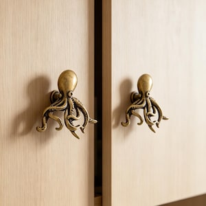 Puede incluir: Dos pomos de armario de metal en tono latón con forma de pulpo, con tentáculos detallados y cabezas redondeadas. Estas piezas de herraje están montadas en puertas de armario de madera clara y presentan un acabado metálico con textura.